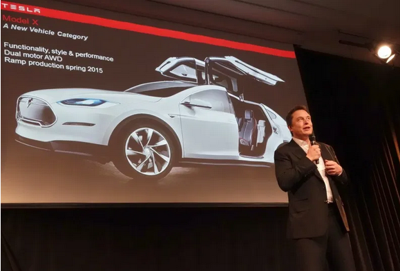 Elon Musk stands in front of the image of a Tesla with a microphone