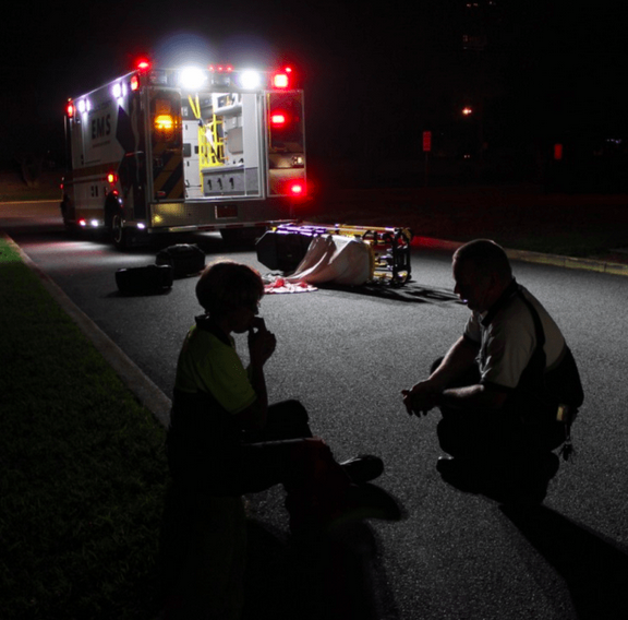 Emergency technician crouches in street at night; emergency vehicle and empty gurney in background