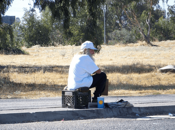 A man with a long white beard sits on a crate in the median of a road