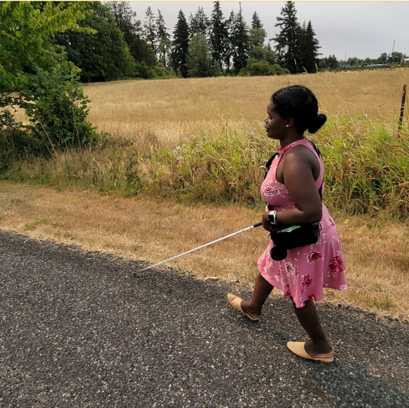 Image description: An African American woman wearing a pink sundress walks along a rural road using a white cane. Via Disability Mobility Initiative