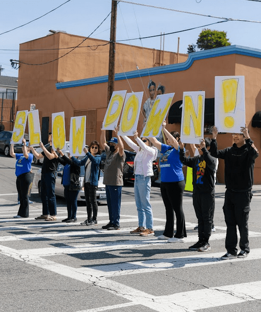 people stand in a line in a crosswalk, holding signs that spell out: SLOW DOWN