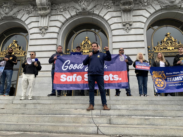 A man speaks into a mic while standing on the steps of city hall, people in the background hold a banner and cheer