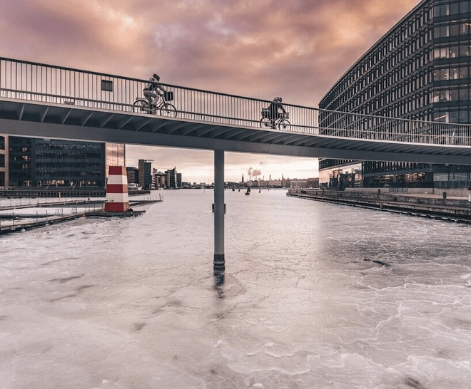 two bike riders cross a bridge stretching over water, with buildings and a cloudy sunset in the background