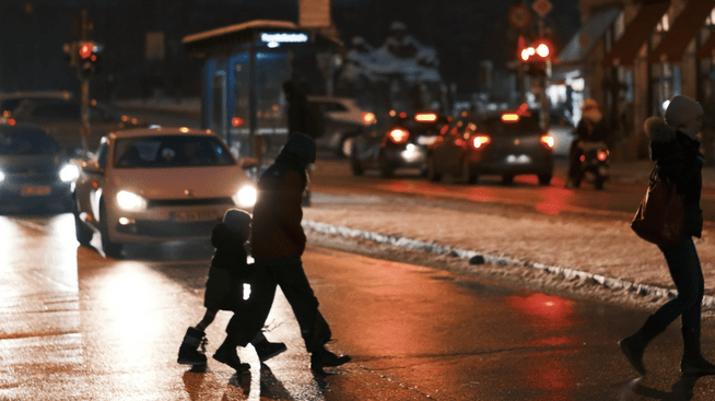 An adult and a child cross a street at night while a car approaches with its headlights on