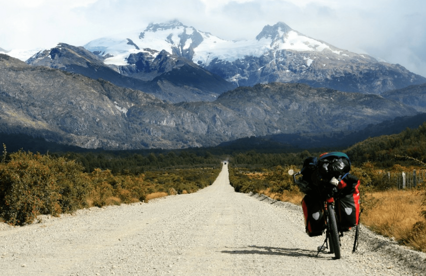 empty dirt road heads into the distance, snow-capped mountains, a heavily laden bike in the foreground
